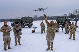 A soldier uses a control as a fellow soldier releases an unmanned aircraft system into the air as others watch from the background in a snowy field.