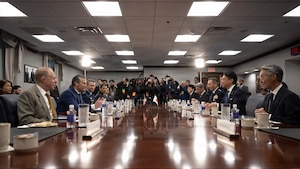 The U.S. secretary of war and Japan's defense minister sit across from each other at a table with other people, as reporters gather at the end.