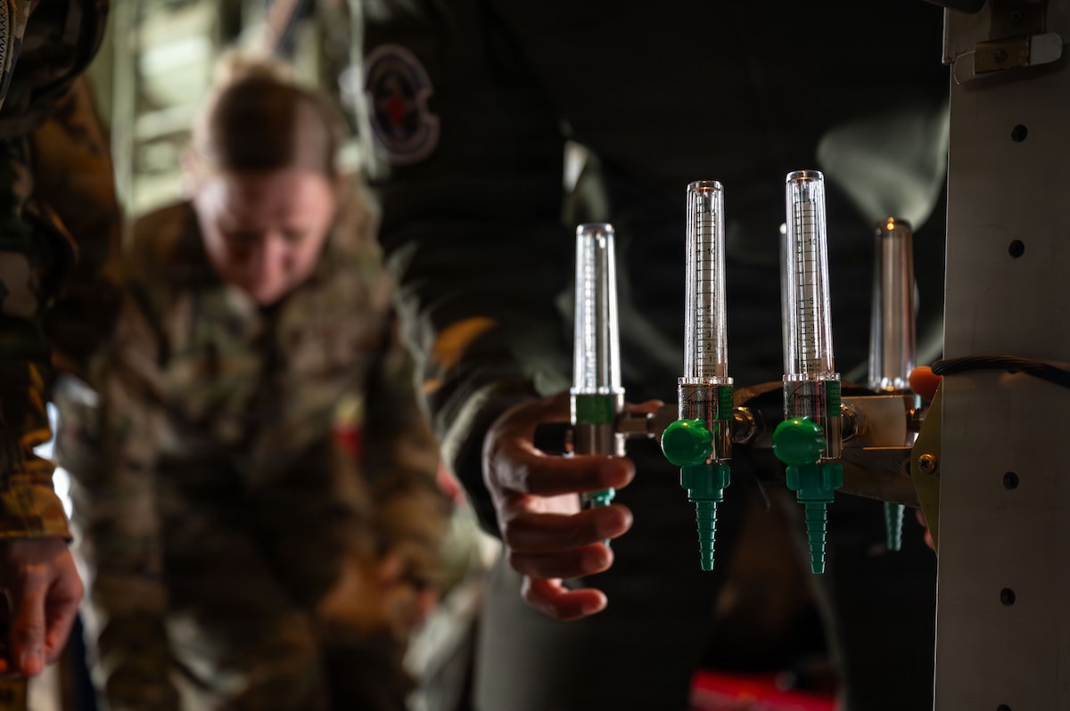 A person in a military fight suit turns the knob on an oxygen regulator. You can see another person in a camouflage military uniform reaching down for something on the floor in the background.