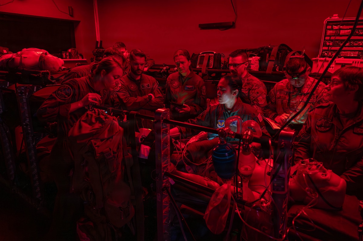 Several service members in flight suits and camouflage military uniforms look at a mannequin on a stretcher. The service members are in a room with medical supplies around them and a red light on that is illuminating the area.