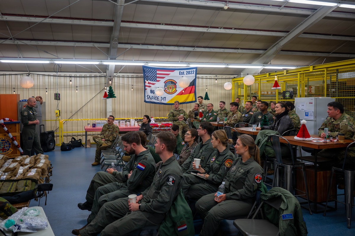 Two dozen service members in flight suits and camouflage military uniforms sit in a room watching another man in a military flight suit speak. There are holiday decorations hanging around the room and a unit flag on the back wall that has the U.S. and German flags incorporated into it.