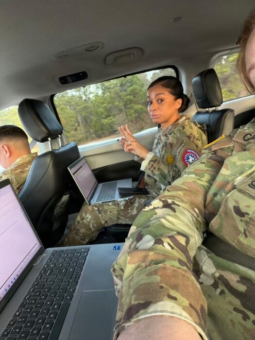 A soldier, wearing a camouflage military uniform, sits in the back seat of a car with a laptop computer in her lap points her fingers up while a soldier sitting next to her in similar dress takes the photo. There is another soldier sitting in the front seat.