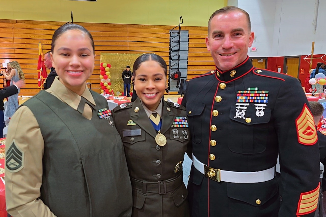 Three service members wearing military dress uniforms smile as they pose for a photo. There are people standing and seated at tables in the background.