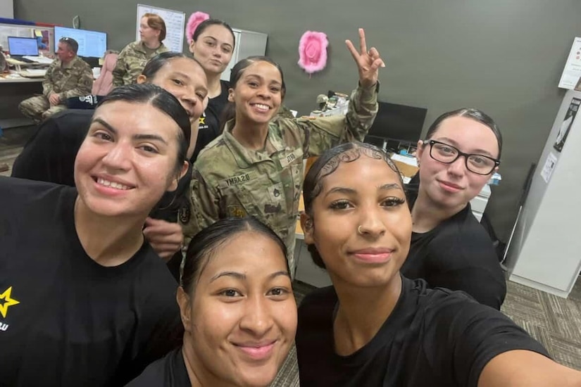 A soldier wearing a camouflage military uniform poses for a photo surrounded by recruits wearing black T-shirts in an office setting. There are two soldiers in the background, one standing and the other sitting in front of a computer.