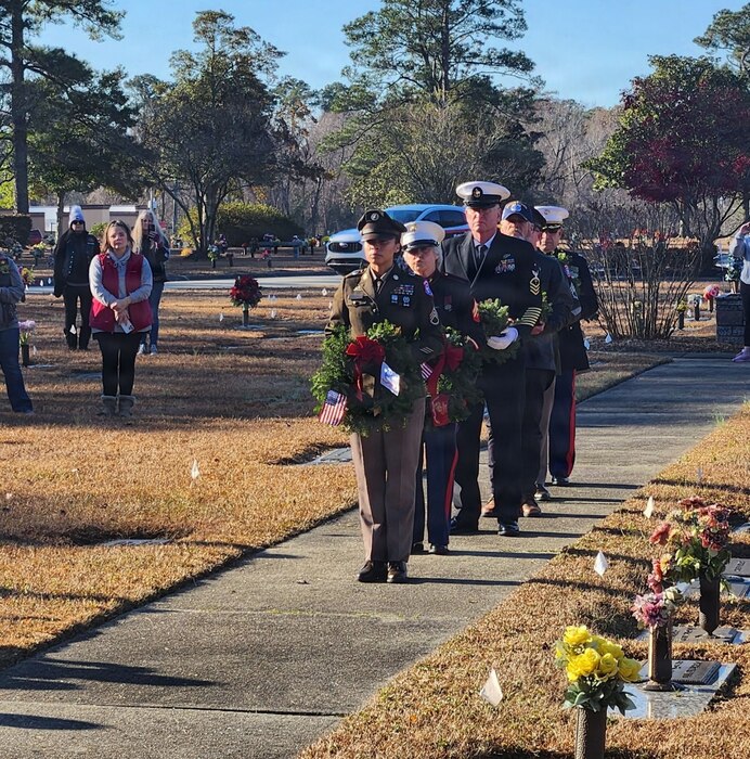 Five service members, each wearing their service’s military dress uniform, stand in a front-facing line while holding wreaths in a cemetery. There are people standing in the background watching the ceremony.