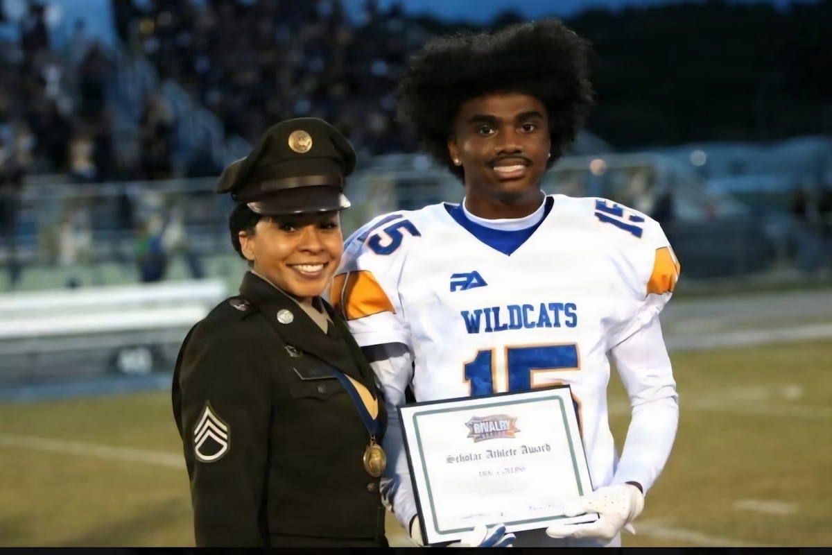 A soldier wearing a military dress uniform poses for a photo with a football player who is holding a plaque.