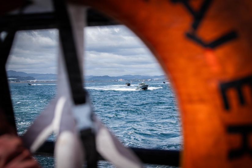 Three small boats sailing in the water on a cloudy day are framed by a life buoy in the foreground. There are mountains in the background.