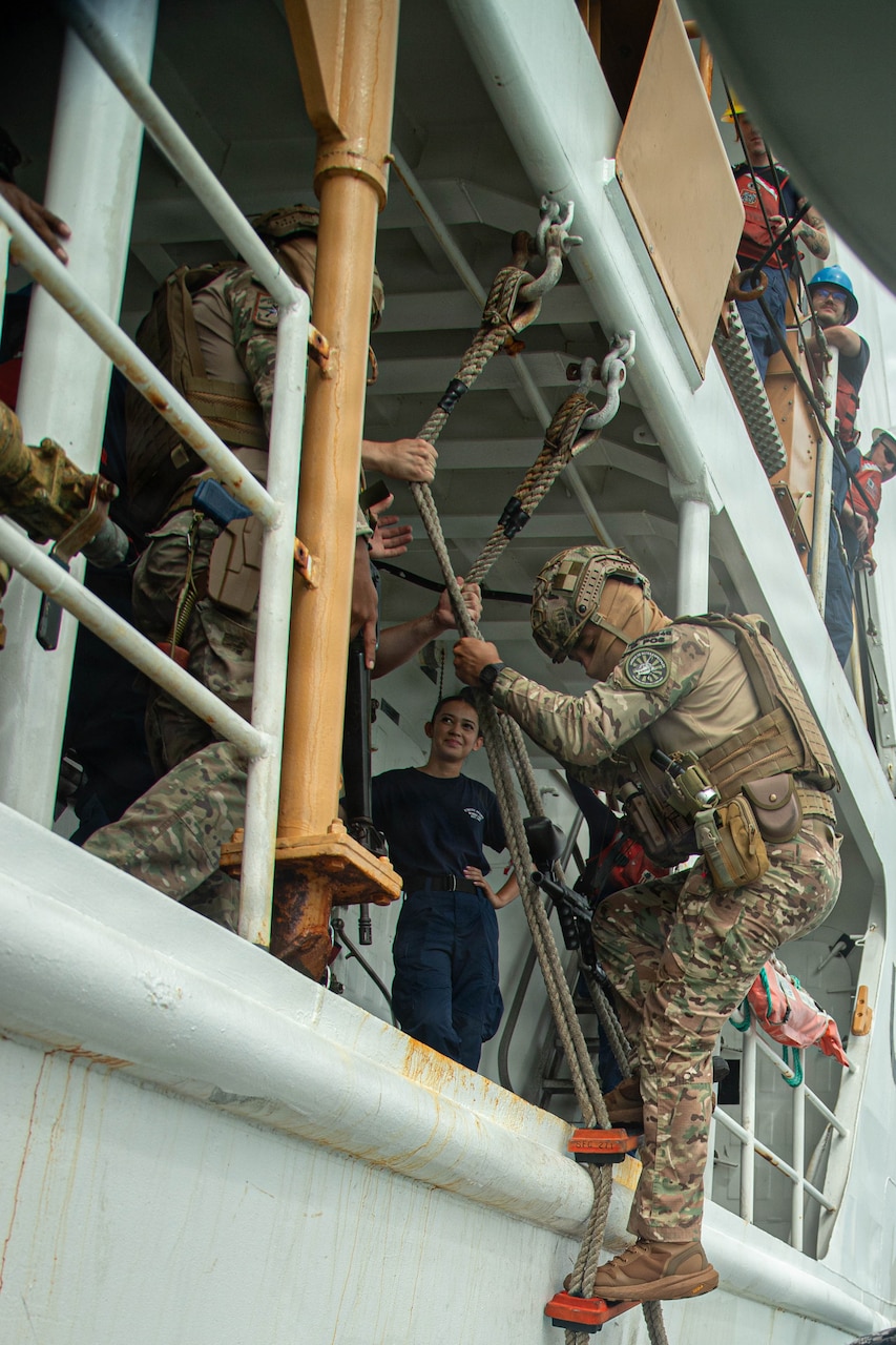 A person in a camouflage military uniform climbs up a rope ladder onto a U.S. Coast Guard boat as six people watch.