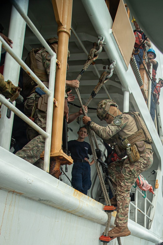 A person in a camouflage military uniform climbs up a rope ladder onto a U.S. Coast Guard boat as six people watch.