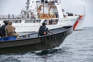 A small boat with three people in camouflage military uniforms sails in the water as a large white boat with U.S. Coast Guard written on the side sails in the background.