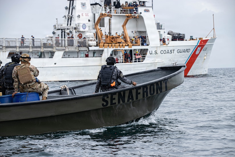 A small boat with three people in camouflage military uniforms sails in the water as a large white boat with U.S. Coast Guard written on the side sails in the background.