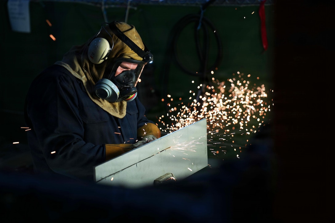 A man wearing a respirator with a face shield, gloves and a protective suit cuts sheet metal, while yellow sparks and light to emit from the object.