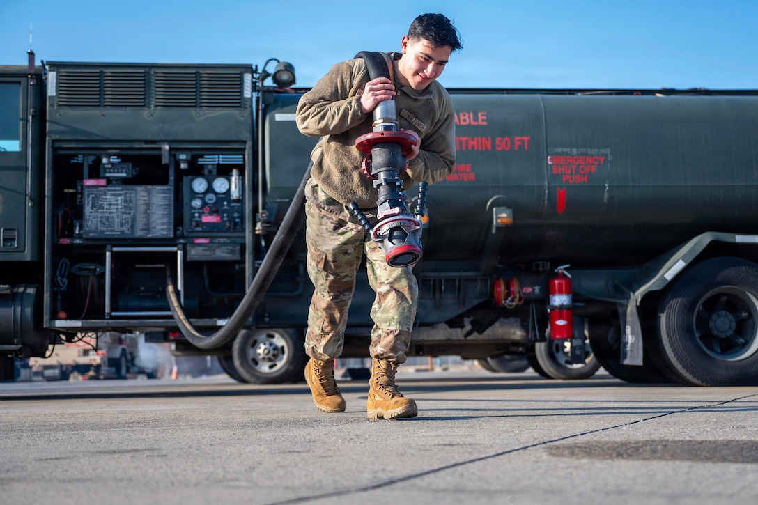 A man wearing a camouflage military uniform pulls a fuel hose from a refueling truck seen in the background as he carries the hose connection over his shoulder.