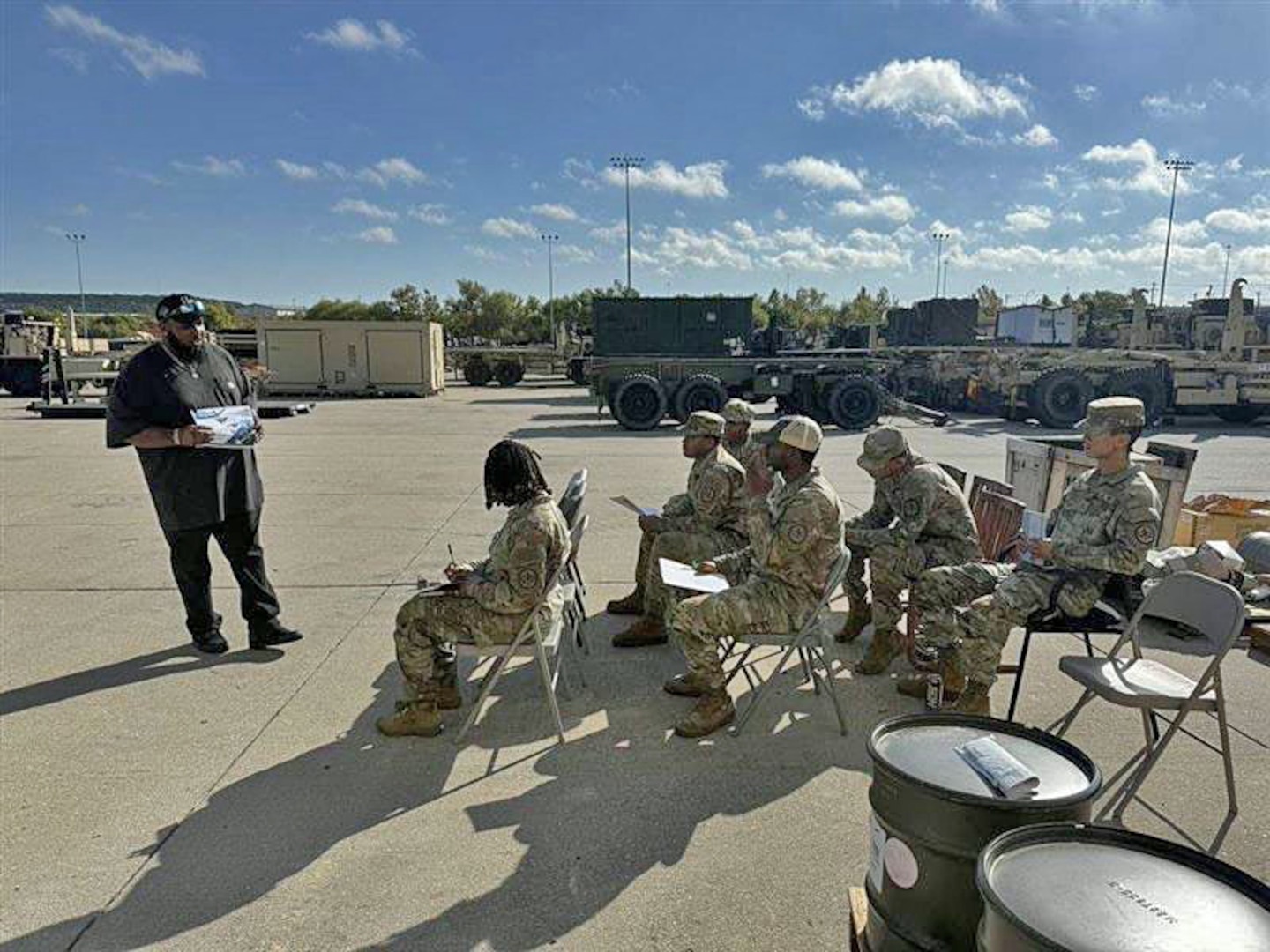 A man on a concrete tarmac speaks to a group of seated soldiers.