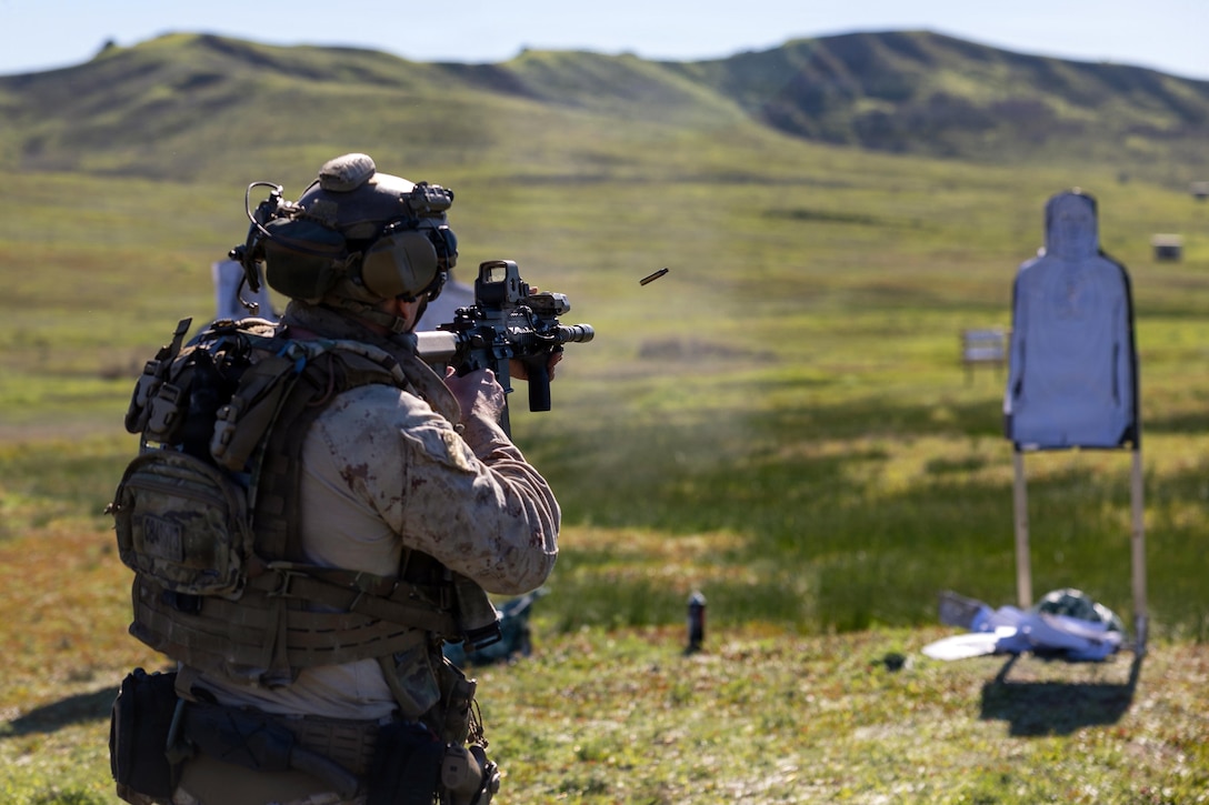 A Marine wearing a camouflage military uniform and combat gear fires a weapon at a target while a spent round flies out at a live-fire range.