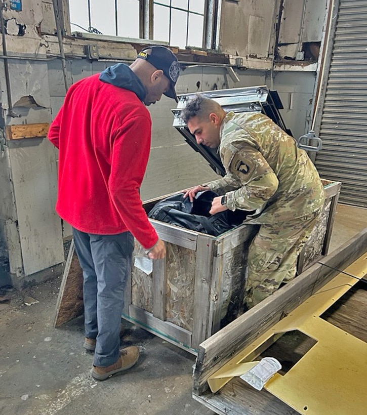 A soldier and civilian look through the contents of a wooden box in a warehouse.