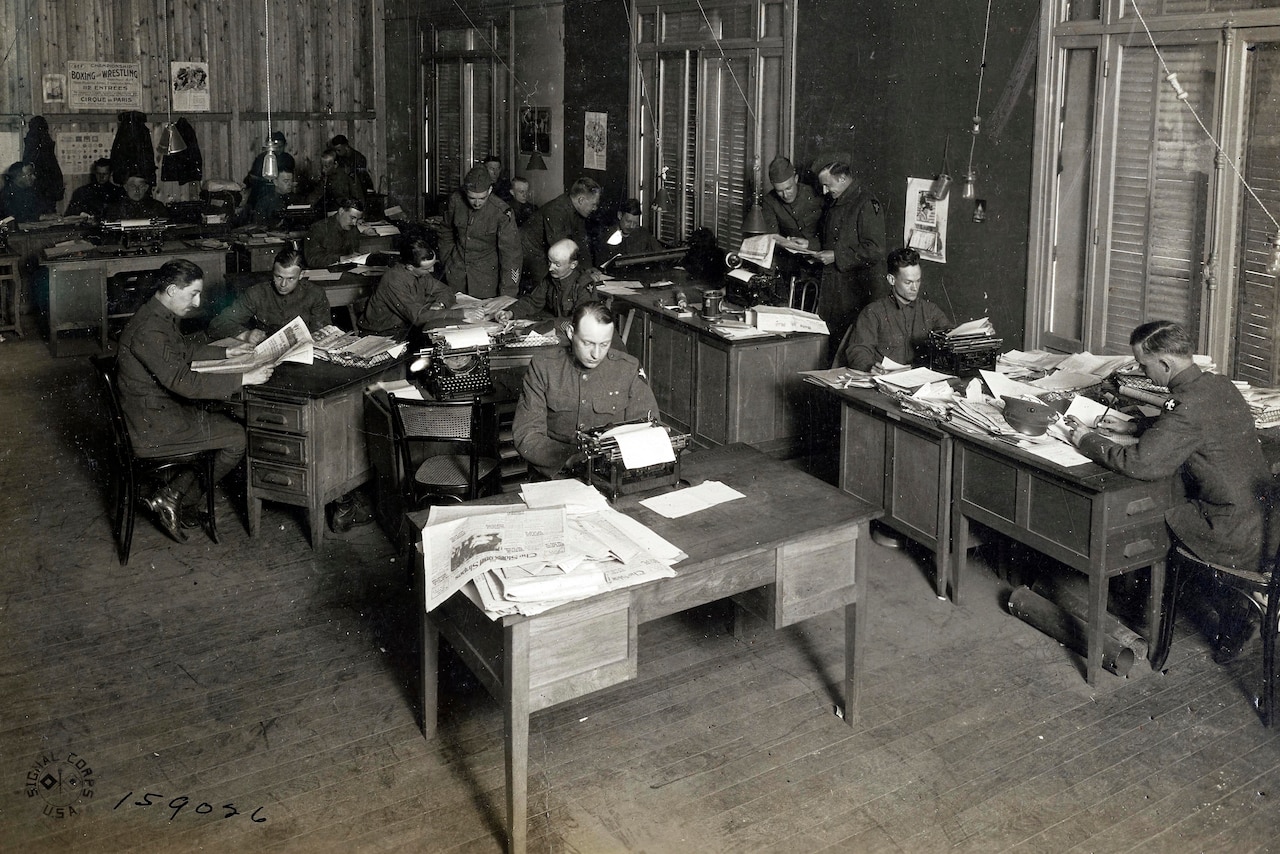 A black and white photo shows more than a dozen men in military uniforms at desks with typewriters.