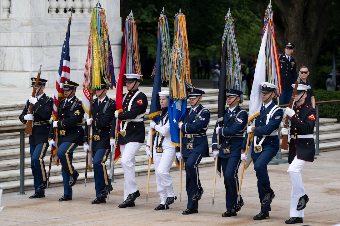 Nine service members wearing official ceremonial military uniforms march in a line next to one another holding flags and rifles outside near stairs.