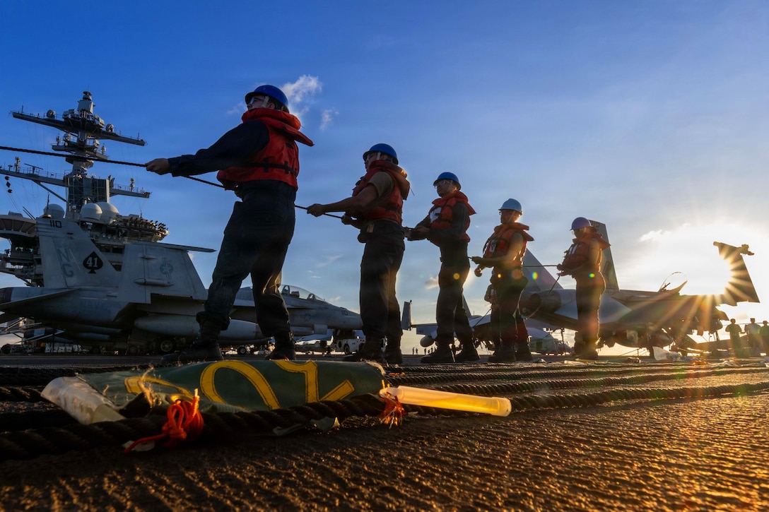 Several sailors wearing hard hats, protective suits and safety vests hold a line while standing in a front-facing line on a flight deck of an aircraft carrier with fighter jets in the background.