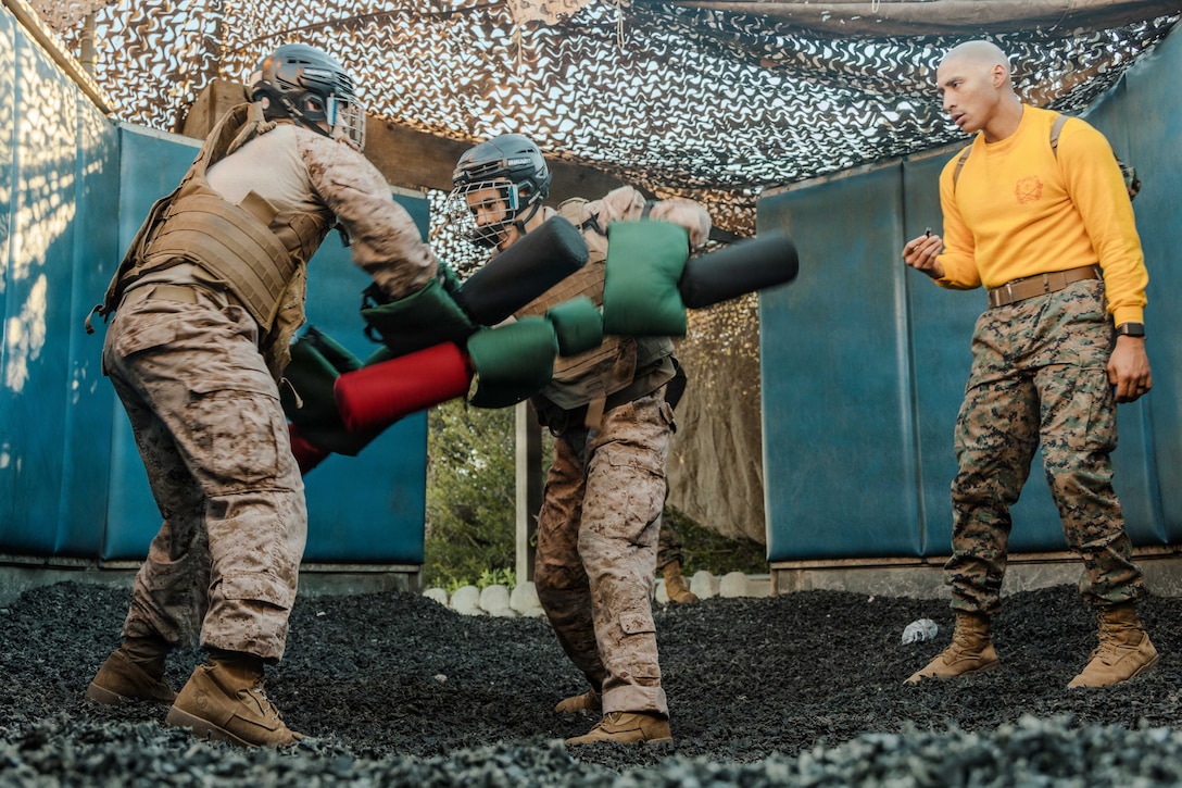 Two Marine Corps recruits wearing headgear and body and hand protection spar with padded sticks in an outdoor setting under netting as a nearby person watches.