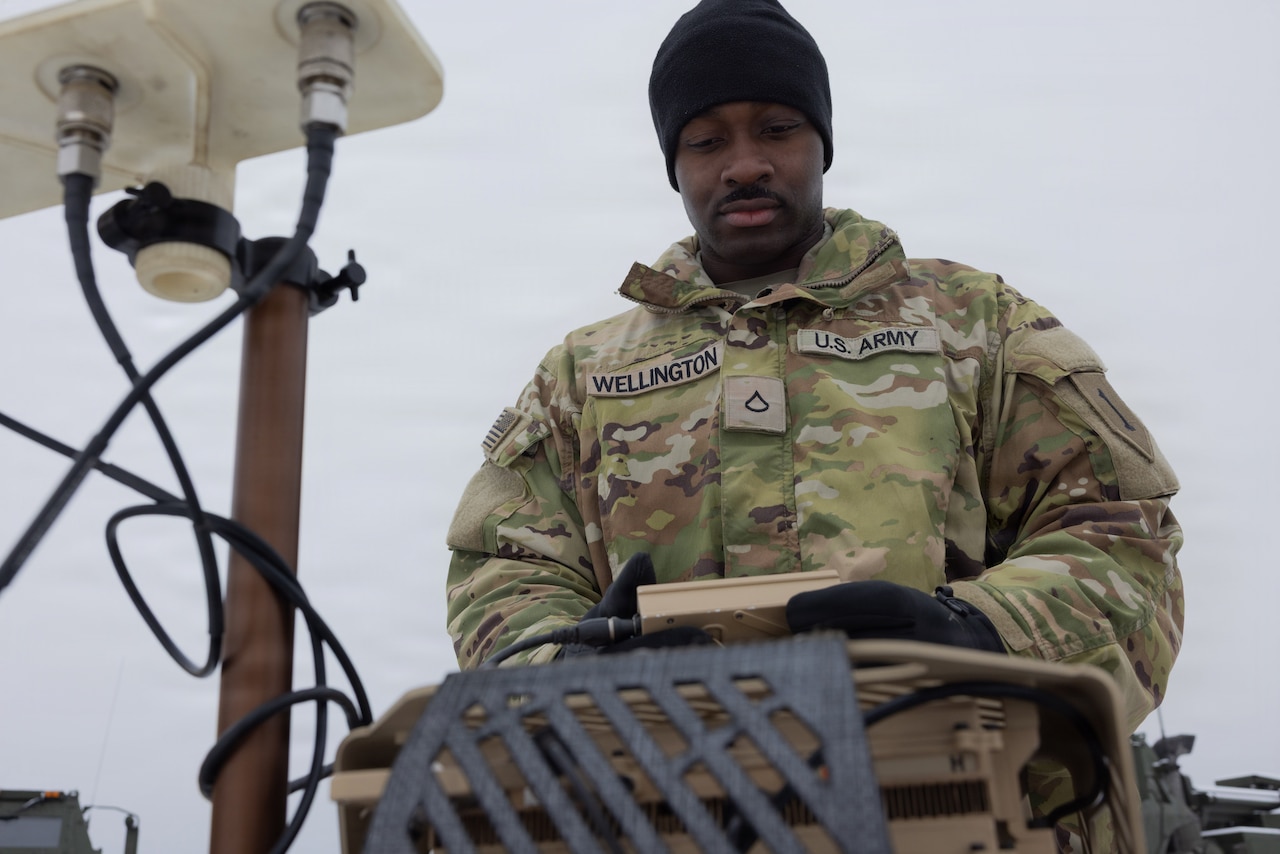 A man wearing winter camouflage military gear looks down at a piece of military equipment.