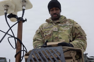 A man wearing winter camouflage military gear looks down at a piece of military equipment.