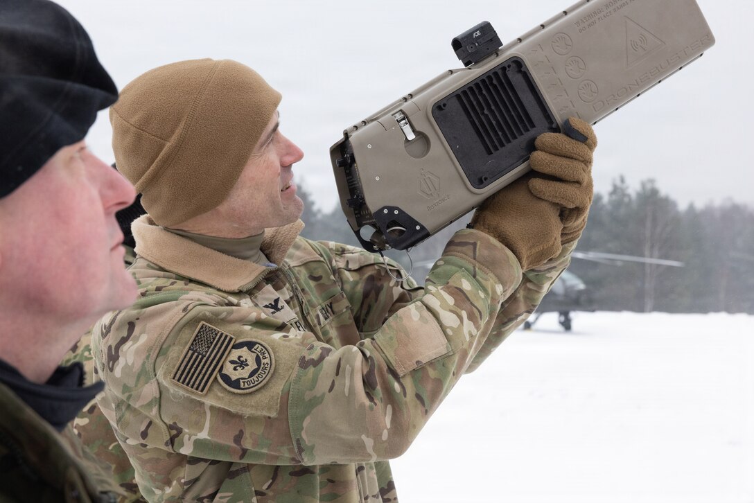 A man in a camouflage military uniform holds a drone busting piece of equipment in his hands as he looks through its viewfinder. There is another man in similar attire standing next to him.