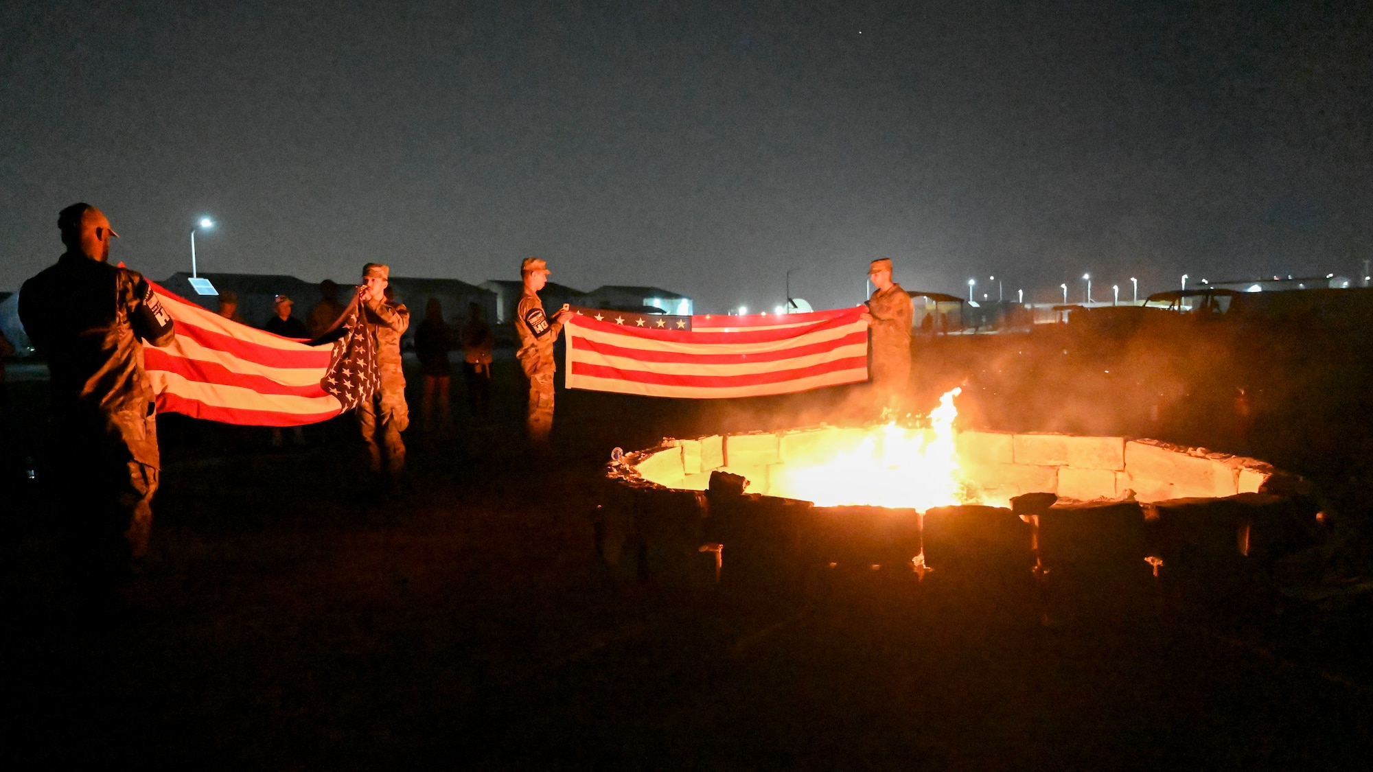 Men in military uniforms fold American flags next to a fire.