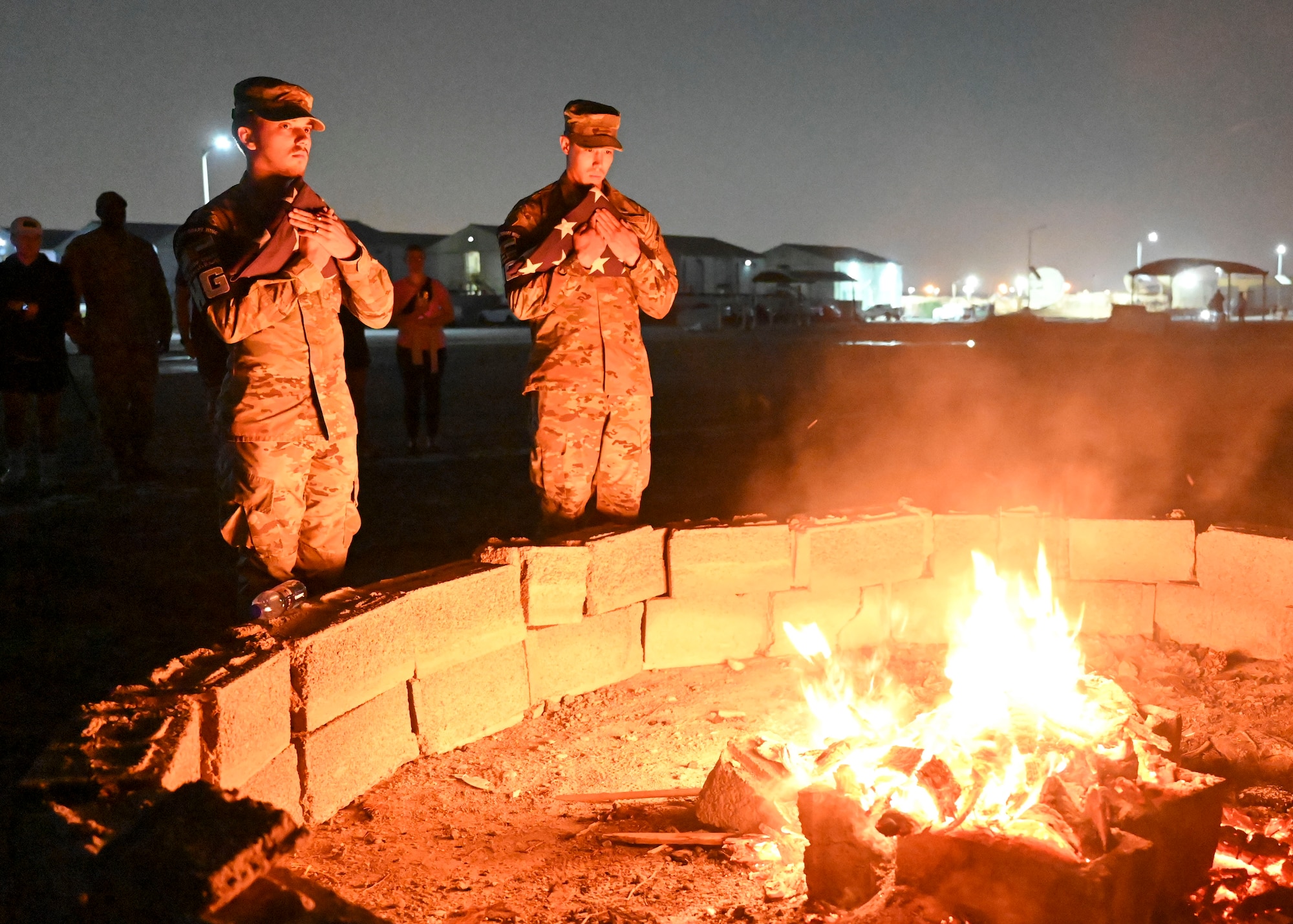 Two men in military uniforms hold American flags next to a fire.