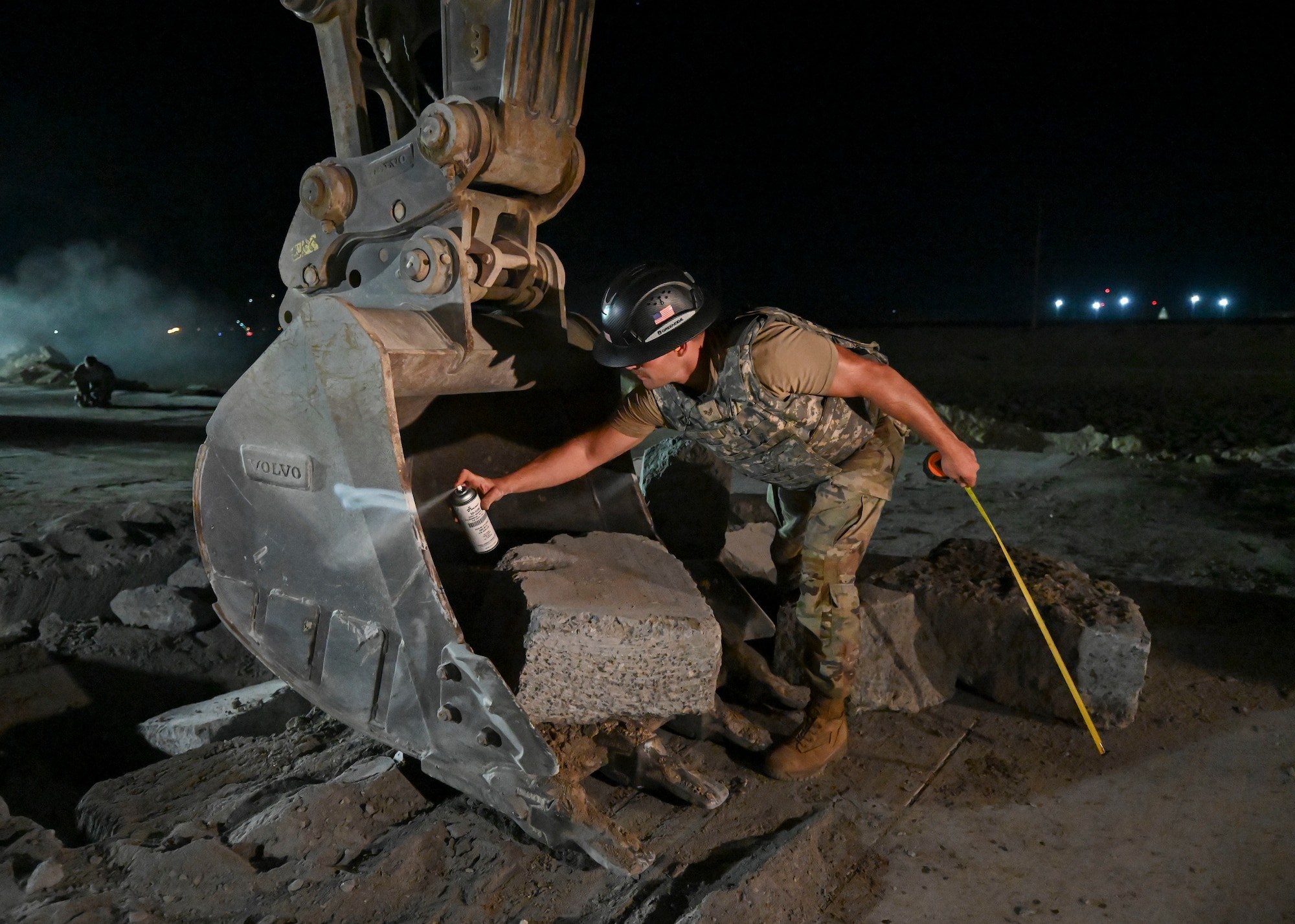 A man in a military uniform uses spray paint to mark a line on a excavator's bucket.