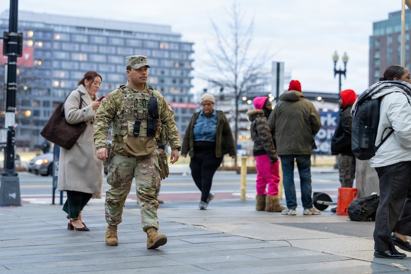 A man in a camouflage military uniform walks down a city street with people in winter attire standing and walking around him