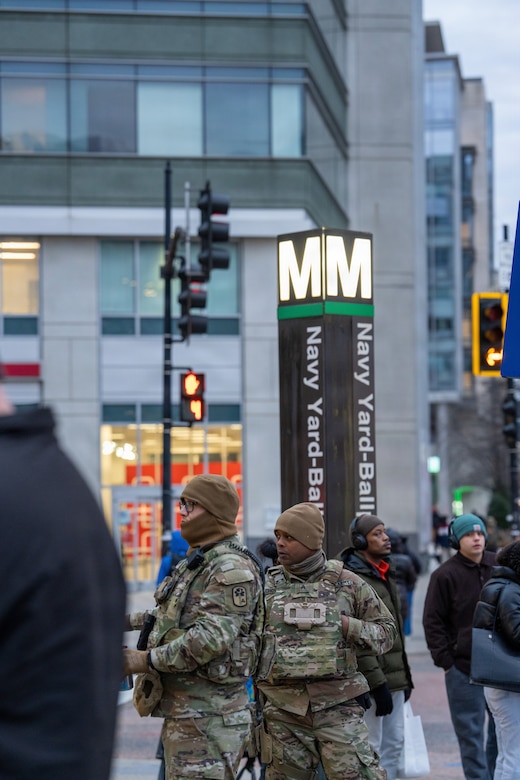 Two men in camouflage military uniforms stand on a street corner with a metro subway station marker behind them. There are several other people in casual winter attire standing around them.