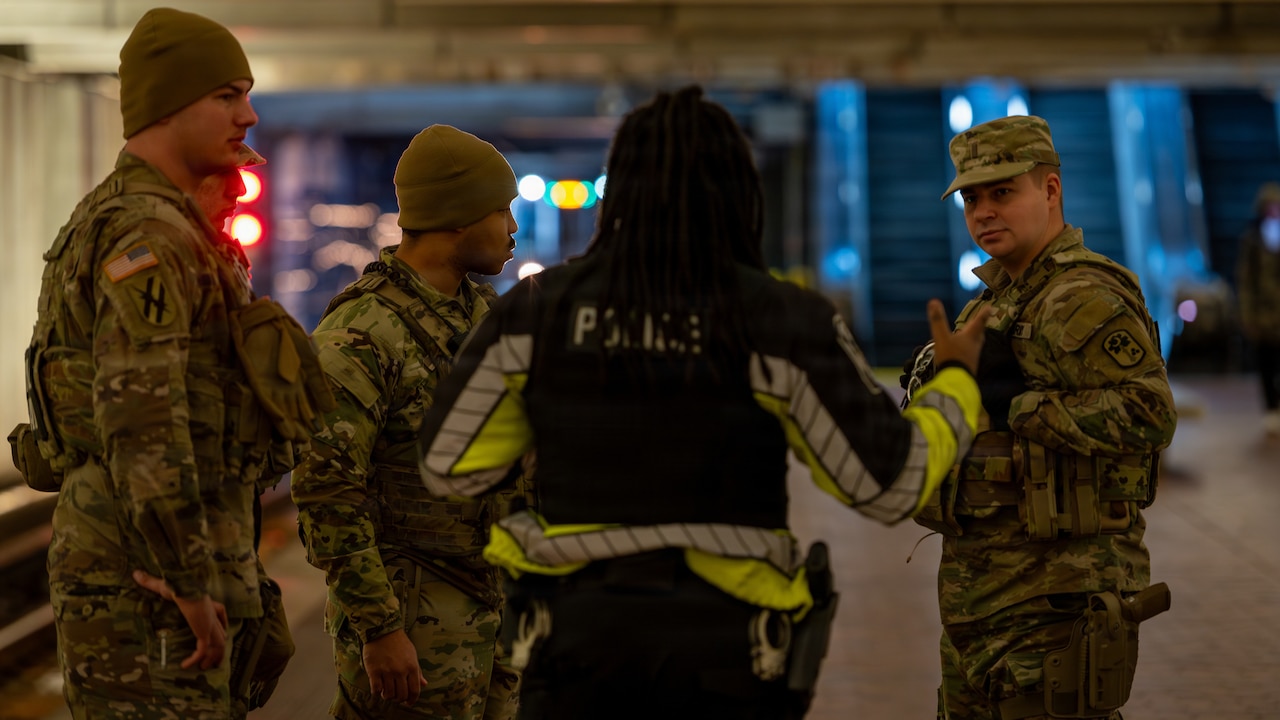 Four men in camouflage military uniforms stand together in an underground subway station as they listen to a man in a police uniform speak to them.