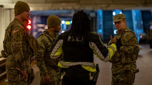 Four men in camouflage military uniforms stand together in an underground subway station as they listen to a man in a police uniform speak to them.