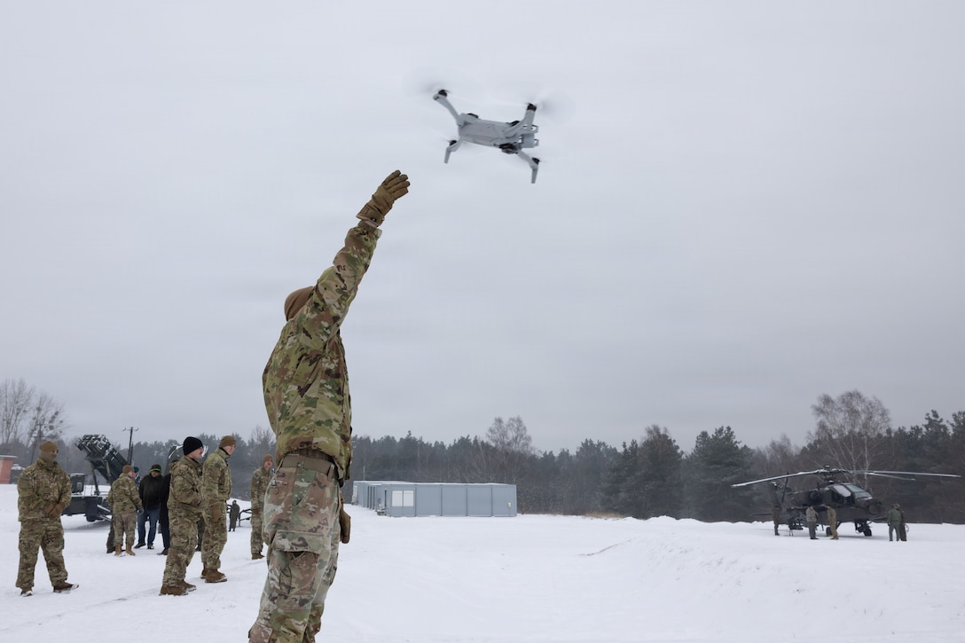 A man in a camouflage military uniform lifts his hand up as he releases a military drone. Other people in similar attire stand in the background.