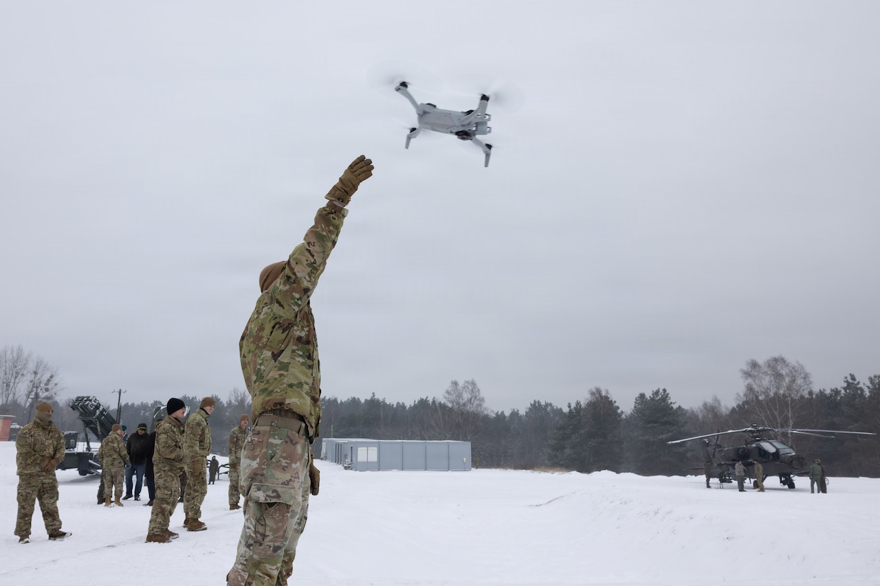 A man in a camouflage military uniform lifts his hand up as he releases a military drone. Other people in similar attire stand in the background.