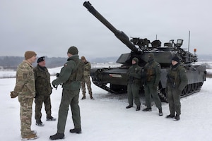 Men in military uniforms stand in front of a military tank talking, while others in similar attire stand next to it.