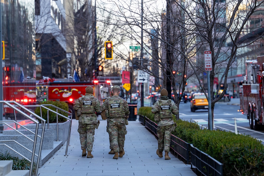 Four people wearing camouflage military uniforms walk down a city street.