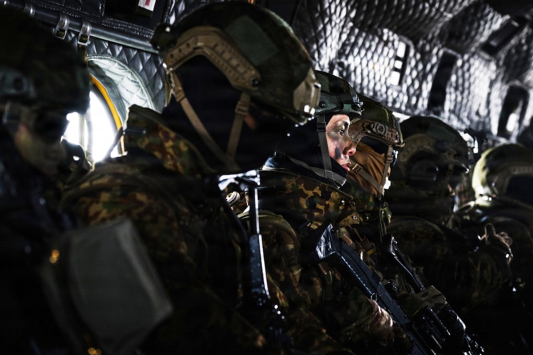 Several people wearing camouflage military uniforms and helmets sit in a row in a dark area.