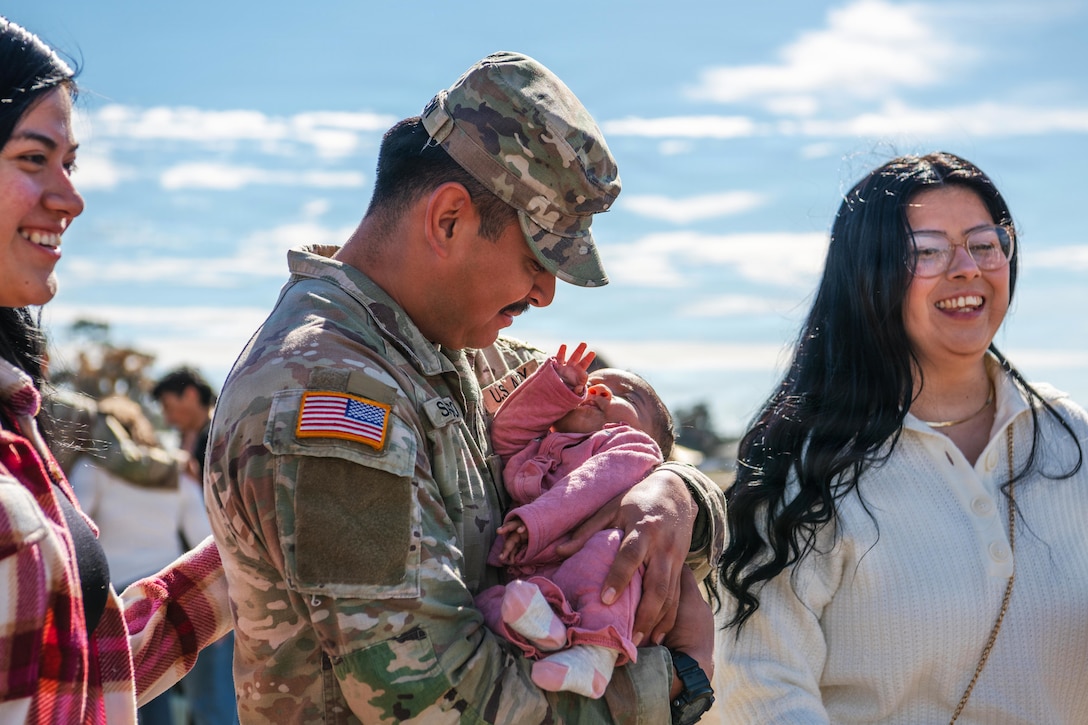 A man wearing a camouflage military uniform holds a small baby dressed in pink, while two women on both sides of him smile and look ahead.
