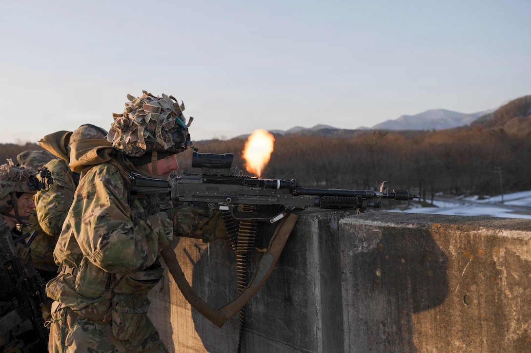 A man wearing a camouflage military uniform and headgear fires a weapon, while people in similar attire do the same outside, with trees and mountains in the background.