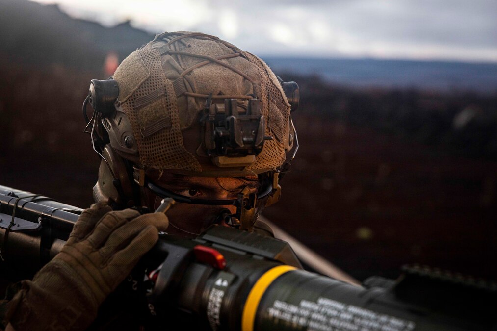 A Marine wearing a helmet and gloves fires a partially visible weapon against a blurred mountainous background.