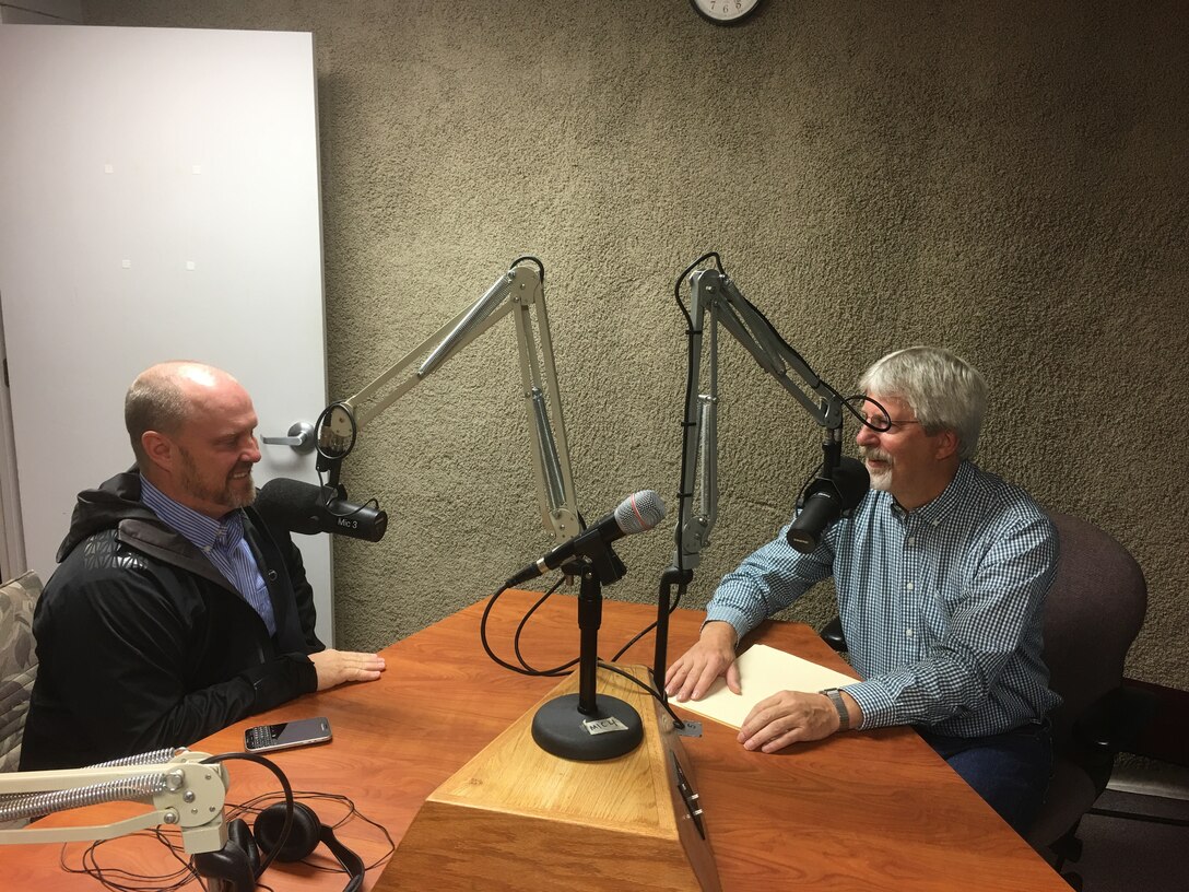 Two men sit at a table with overhead microphones for an interview.