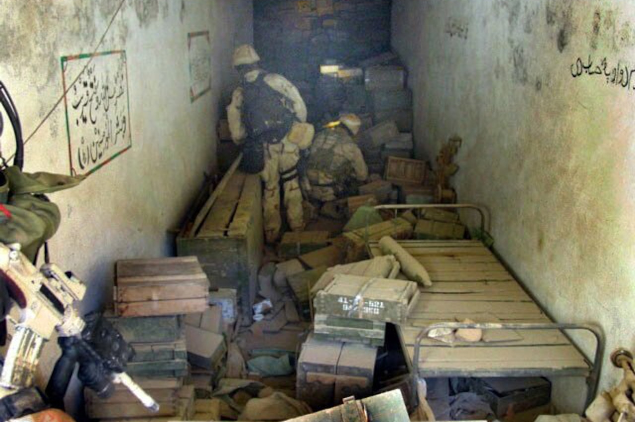 Two men in military camouflage uniforms look through a dingy concrete room filled with cots and boxes. Arabic writing is scrawled on the walls.