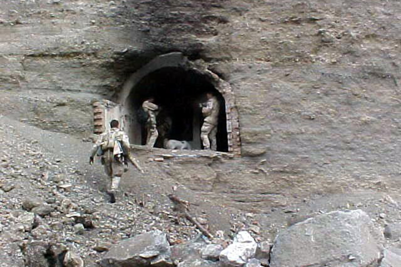 Three men in tactical military gear stand in the entrance to a rock cave.