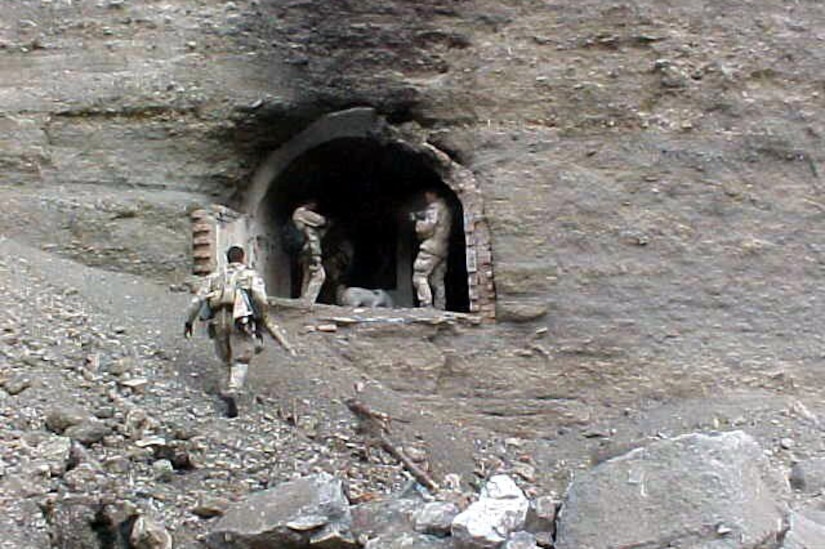 Three men in tactical military gear stand in the entrance to a rock cave.