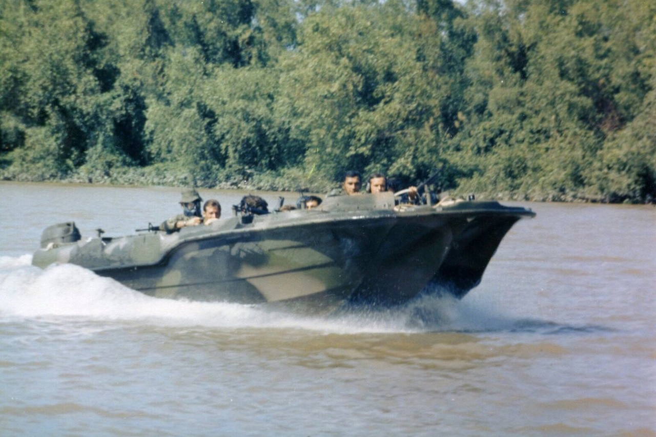 Men in a camouflaged speed boat crouch low as they speed through a river.