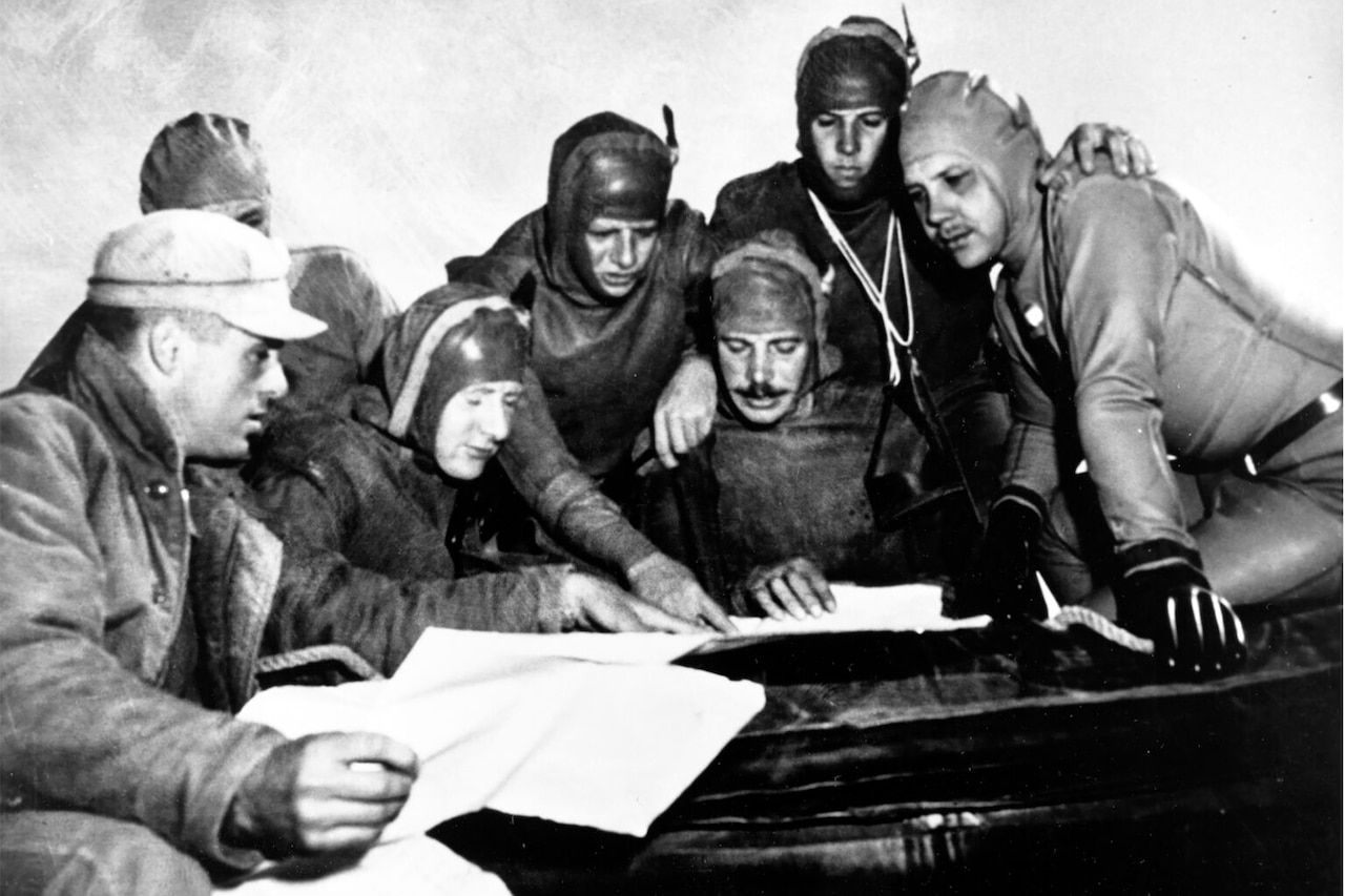 Seven men, most of whom are in wetsuits and swim caps, gather together to look at a bunch of papers.