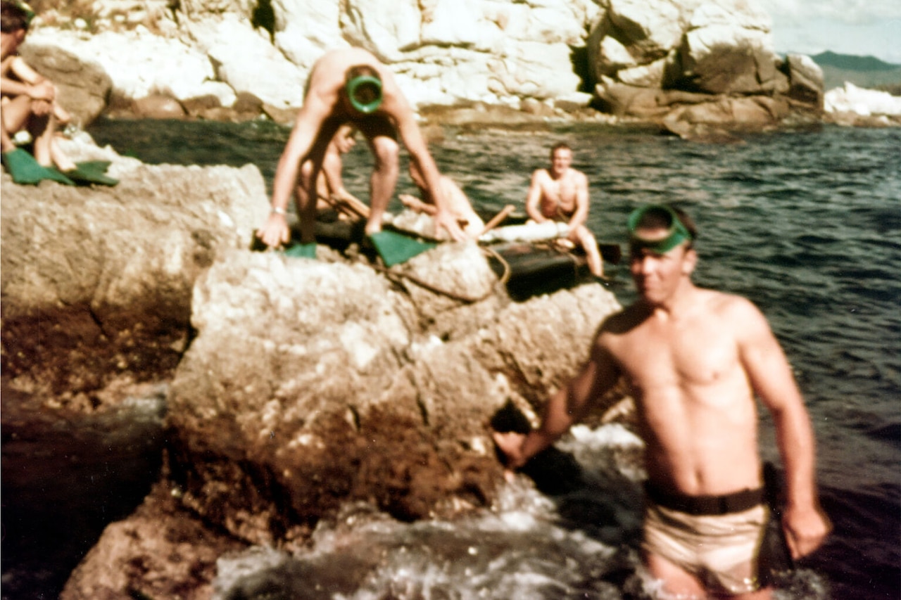 Five men in swim shorts and snorkels climb over rocks jutting into the ocean.