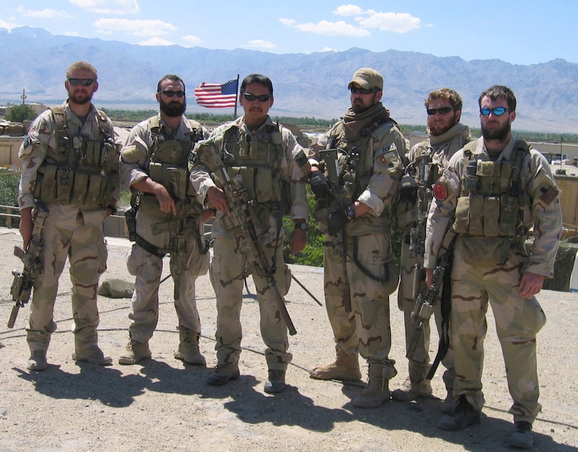 Six men wearing tactical camouflage military gear and carrying automatic weapons pose for a group photo in the desert. Mountains and an American flag fly behind them.